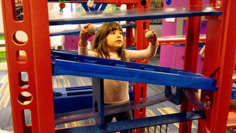 a young girl stands among several sets of towers and ramps, putting balls into the ramps