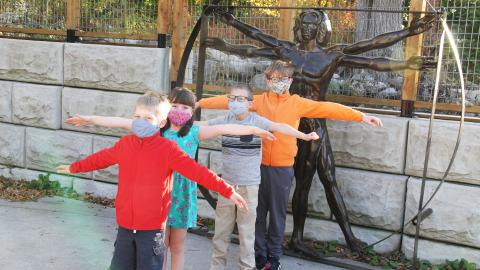 four children stand in front of a metal version of da Vinci's Vitruvian Man, with their arms and legs spread