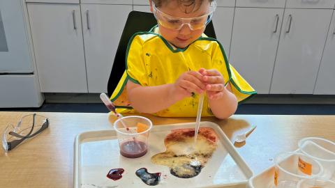 A small boy wearing a smock and safety goggles uses a pipette with various liquids of different colors on a tray.